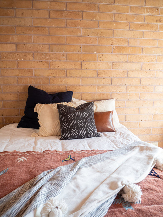 black mudcloth cushion sitting on a bed with various shaped and coloured cushions behind and a throw coming into shot from the bottom 